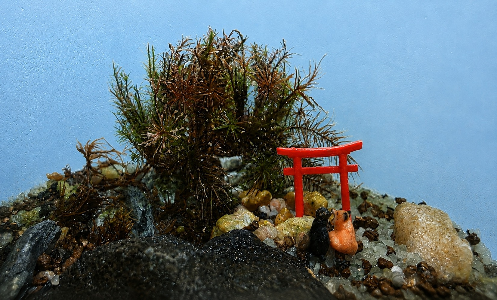 A miniature moss garden with a red torii gate and tiny fox figures against a blue sky background